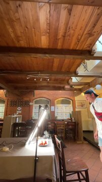 man stands on wooden chair and leans over table to operate camera rig while bright led tube lights illuminate plate of red food inside rustic cafe with exposed