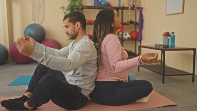 Man and pregnant woman sit back to back on yoga mat holding meditation mudra in a studio; calm bonding.