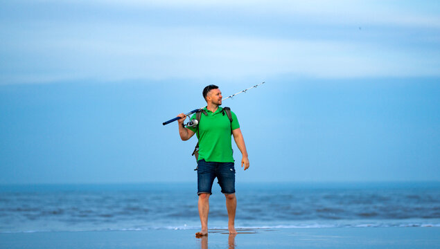 Fishing man walks near ocean water. Fishing rod and reel in hand. Fishing on shoreline with surf waves. Fishing lifestyle shows outdoor recreation.