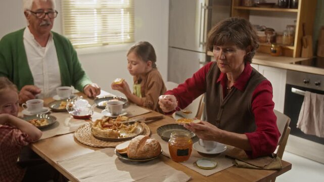 Senior Caucasian grandmother spreading jam on bread and giving it to granddaughter during breakfast with family. Footage suitable for generational lifestyle campaigns and food advertising