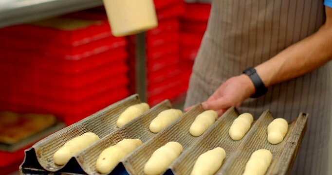 close-up view of baker at chest height misting and steadying rows of raw baguette-shaped dough on ribbed metal proofing tray inside busy bakery, worker wears, with