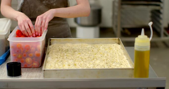 hands of baker in flour-dusted apron placing halved cherry tomatoes into dimpled focaccia dough on rectangular metal tray at stainless steel workstation, plastic