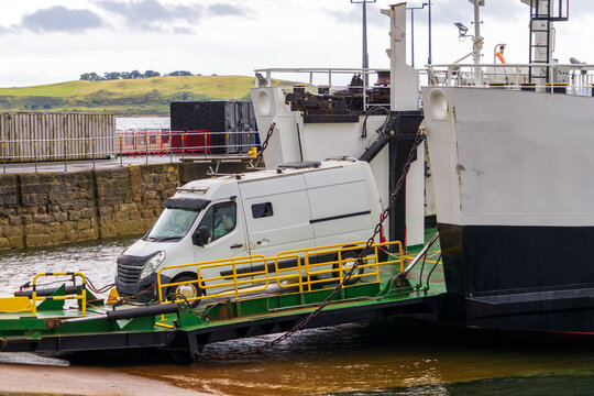 A white camper van driving onto the ramp of  roll-on/roll-off ferry, leaves open car deck via hydraulic  bow ramp at a coastal slipway. A modern double-ended hybrid ferry near a stone pier. 