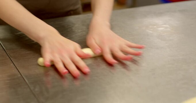 close-up of baker hands pressing and rolling small cylindrical dough piece on scratched stainless steel countertop, partial forearms and torso visible with blurred