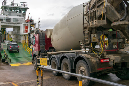 A heavy-duty concrete mixer truck loaded onto a roll-on/roll-off ferry. Multi-axle rigid chassis with a rotating mixing drum. Parked on the vehicle deck of a ferry next to a passenger car. 