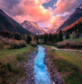 Aerial view of a blue mountain stream in green valley with autumn forests and snow-capped peaks at sunset. Top view of river with blue water, rocks in snow, trees and colorful cloudy sky in fall. Alps