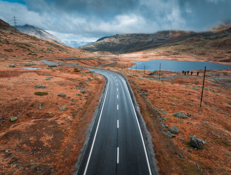 Aerial drone view of wet winding road in the Swiss Alps, Switzerland, with alpine lake, dramatic clouds, golden slopes and rocky terrain on a cold moody autumn day. Top view of mountain alpine road