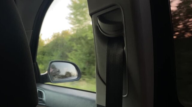 passenger-side interior detail showing textured grey b-pillar and black seatbelt retractor in sharp focus while side mirror and outside foliage shift past window