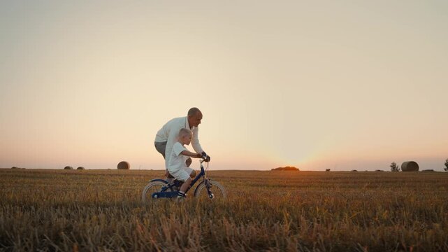 Grandfather guiding young child learning to ride bicycle through golden countryside field at tranquil sunset, sharing love, support, and joyful outdoor exploration