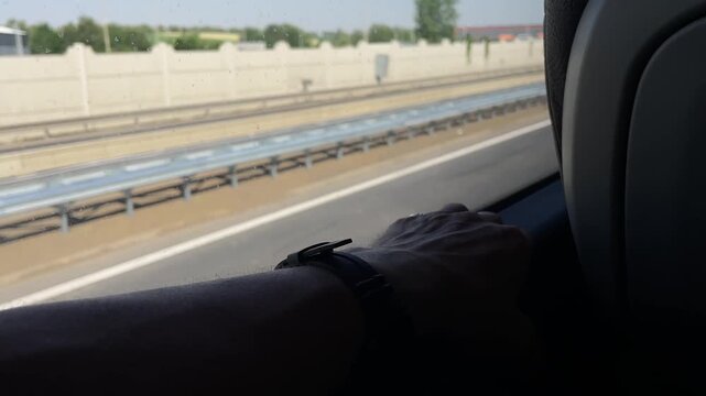 close-up passenger-side view of forearm and hand resting on interior window ledge with wristwatch strap and ring visible as highway, guardrail, and open fields hand