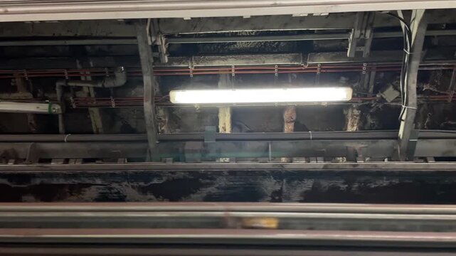 low-angle motion view showing exposed industrial ceiling with bright fluorescent tube, metal support beams, cable trays, and parallel orange electrical cables beam