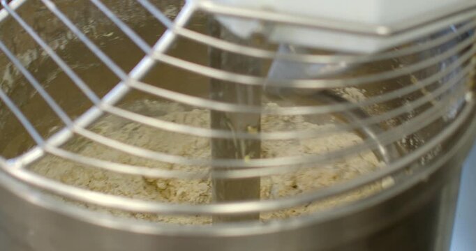 close-up view of industrial spiral mixer mixing dry flour into dough inside polished stainless steel bowl, seen through horizontal safety cage bars, central, with