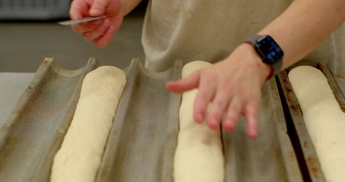 close-up motion scene of baker's hands scoring and steadying baguette dough placed in corrugated mesh proofing tray on stainless workbench, visible action shows