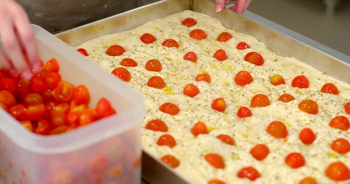 close-up of hands topping pre-dimpled focaccia dough with halved cherry tomatoes in large metal sheet pan, translucent plastic container of tomato halves at left