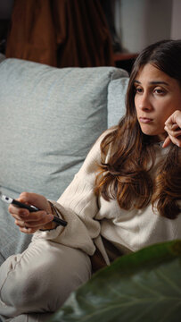 Young Latina woman sitting bored on a sofa while watching television, capturing idle time, home entertainment, media consumption, and everyday leisure.