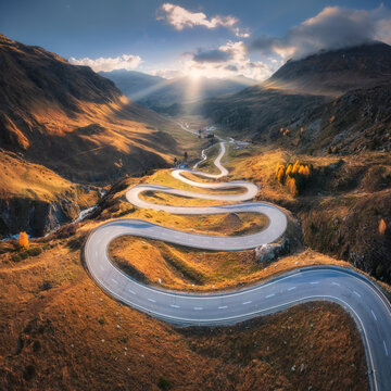 Aerial view of winding mountain road in the Swiss Alps, Switzerland, with sharp curves, golden slopes, valley and dramatic sun rays breaking through clouds over alpine peaks. Top drone view of road