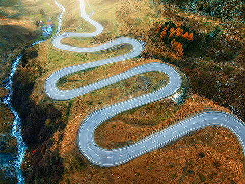 Aerial view of winding mountain road, river in alpine valley at sunset in autumn. Top drone view of road in Swiss mountain pass, yellow grass, trees in fall. Colorful landscape. Alps in Switzerland
