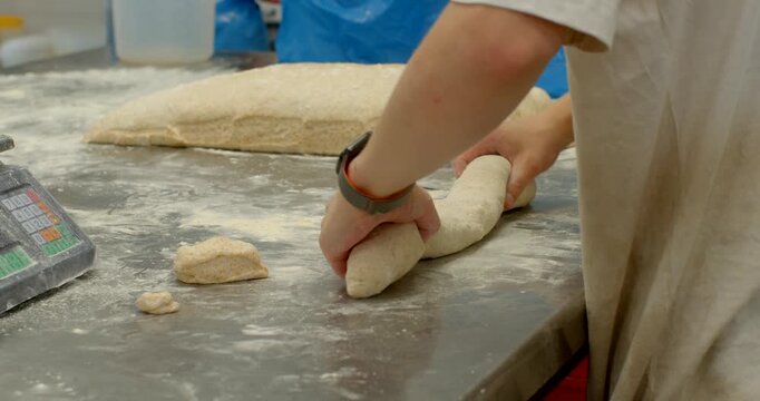 close-up motion of baker hands shaping and portioning dough on floured stainless steel worktable, scene shows long proofed dough mass and smaller cut portions bakery