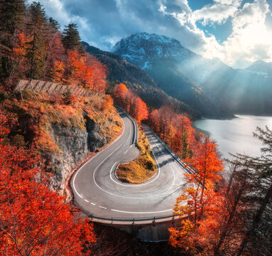 Aerial view of winding mountain road at sunset in autumn. Alps, Italy. Curved alpine road, snowy rocks, trees, red leaves, lake, sky with sunbeams. Colorful. Top view of road on mountain pass in fall