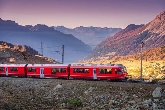 Red Bernina Express train traveling through a high alpine valley in the Swiss Alps, Switzerland, with rocky slopes, pine trees and layered mountain silhouettes in soft evening light at sunset. Autumn