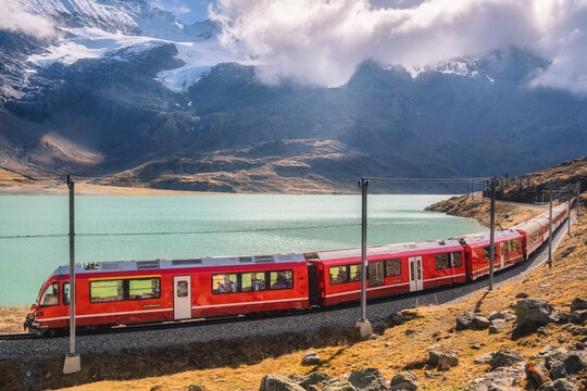 Red train is moving along railway in swiss alps mountain range near lake on a sunny day with blue sky in autumn. Landscape. Bernina Express train, orange grass, snowy rocks at sunset in fall. Travel