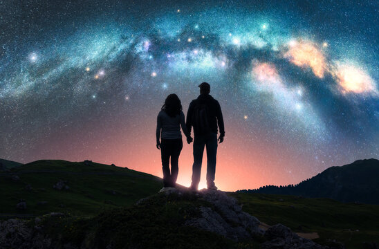 Couple of hikers holding hands standing on a rocky outcrop in mountains, under pink night starry sky with glowing Milky Way. Landscape with silhouettes of man and woman, bright stars and milky way