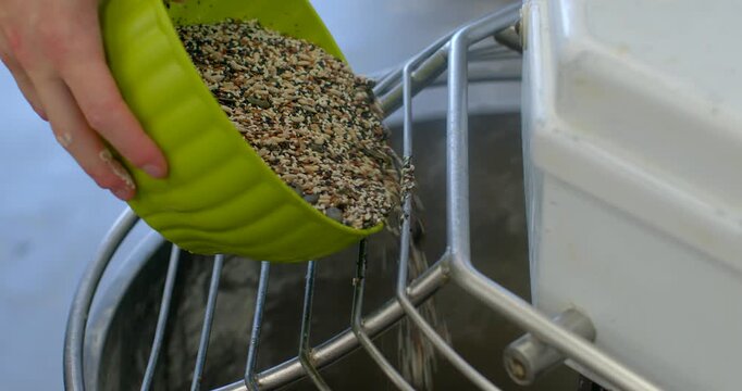 close-up of hand tipping green silicone bowl to pour mixed seeds over stainless steel safety guard into industrial mixer, visible seed mix includes light and dark