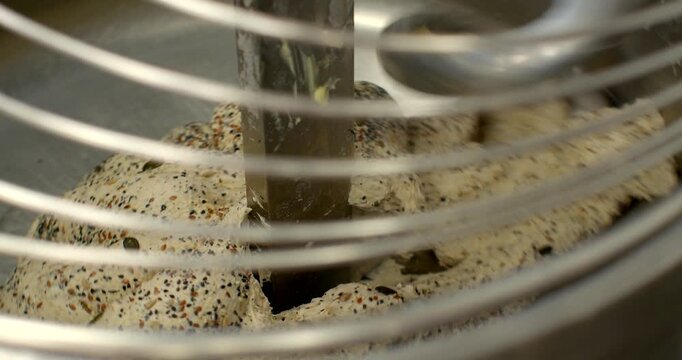 close-up of seeded bread dough being folded and compressed around central metal mixing blade inside stainless steel bowl, circular wire safety guard bars cross dough