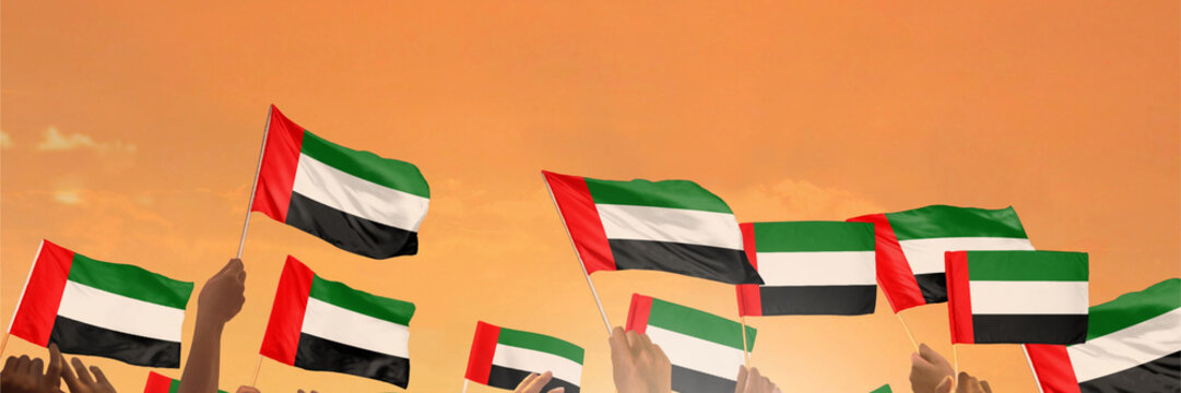 Hands wave UAE flags against an orange sky during a celebration.