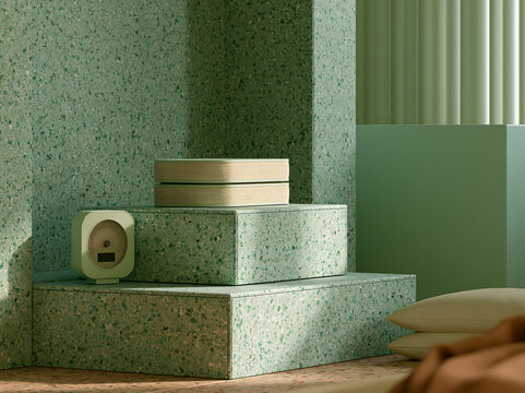 A small square digital clock and stacked rectangular boxes on a green terrazzo platform in a minimalist room with soft light and fabric textures