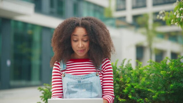 African American female with curly hair wearing red and jeans smiling while typing on laptop. Thinking about best reply and enjoying fresh air working outdoors near greenery.