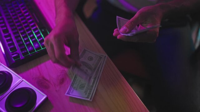 Man counting cash with hands on wooden desk near gaming keyboard and speakers under purple studio lighting; concentration hustle earnings.
