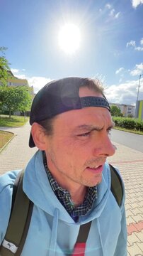 candid close view of man walking along suburban sidewalk, squinting and turning head toward intense midday sun, crisp shadows and bright highlights in neighborhood