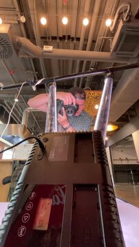 photographer in baseball cap and plaid shirt lowers dslr from eye level behind transparent supports inside industrial-style interior, foreground dominated by, with