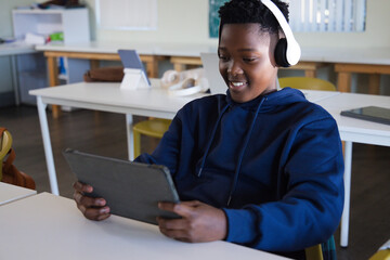 Teen sitting at school desk holding tablet in landscape wearing navy hoodie, white headphones © WavebreakMediaMicro