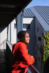 Obraz premium African American youth male wearing red hoodie leaning on balcony railing, smiling, looking right