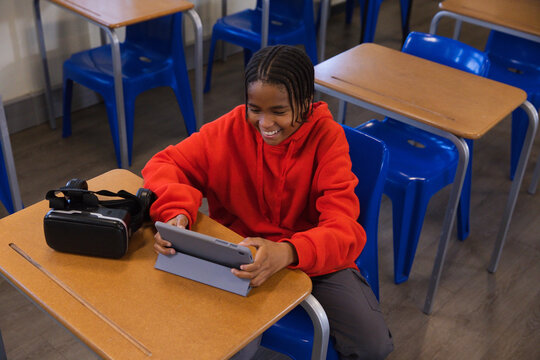 African youth sitting at school desk wearing red hoodie holding tablet on folio VR headset nearby