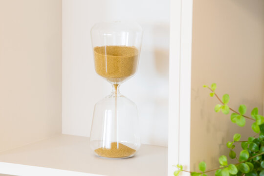 Fine golden sand passes through a glass hourglass placed on a minimalist shelf. A green plant in the background contrasts with the neutral white surroundings.