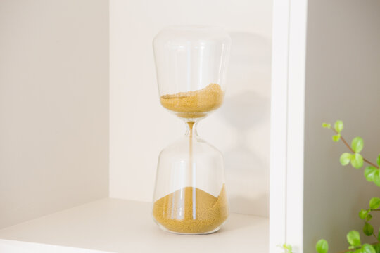 A transparent hourglass measures time with fine golden sand flowing between its chambers. The simple glass object is positioned on a white built-in shelf.