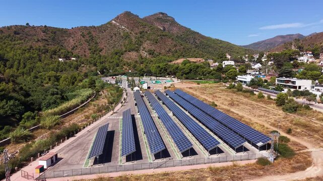 Large solar carport installation with multiple rows of photovoltaic panels in a sunny Mediterranean valley, surrounded by green mountains and a small Spanish hillside village