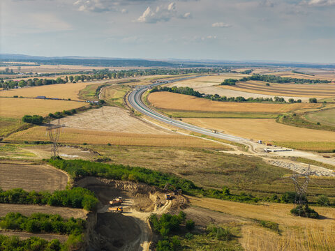 Aerial view of the Autostrada A7 highway construction near Bacau, showing expansive agricultural fields, excavators, earthworks, and sunny countryside landscape. Moldova Highway in Romania