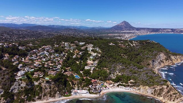 Aerial view of Javea, Spain, showing luxury villas nestled among pine forests on rocky cliffs above turquoise Mediterranean coves with Montgo mountain backdrop