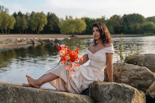Woman sitting on rocks by water holding a bouquet of flowers in a park during sunset Concept of relaxation, freedom, happiness, wellness and connection with nature 