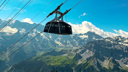 Cable Car Crossing Snowy Alpine Mountain Landscape. Cable car crosses above green alpine slopes with rugged snowy mountains beneath a bright blue sky and white clouds. © Iryna
