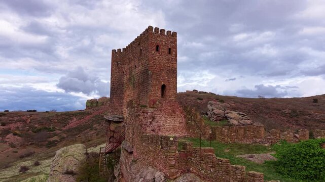 Ancient red stone tower of Zafra Castle rising above rocky hillside in Guadalajara province, Spain, under dramatic cloudy sky with surrounding medieval walls