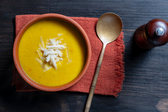 Yellow autumn cream soup served in ceramic bowl with grated cheese toppings, vintage spoon and pepper mill on red napkin, closeup. Ideal for food blogging, restaurant menus and comfort food concepts