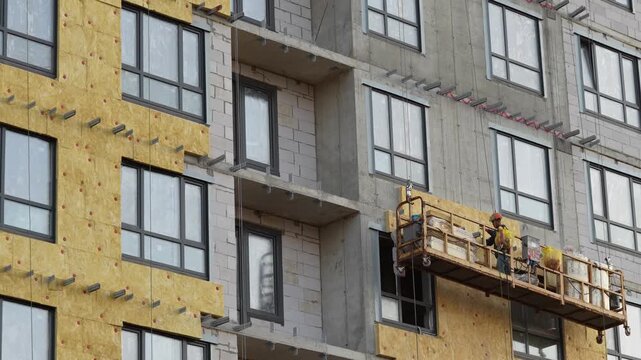 Construction workers installing thermal insulation panels on a multistory building facade from a suspended platform, showing energy efficiency upgrades and modern exterior construction