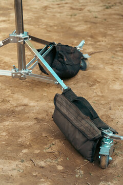A professional metal light stand base equipped with caster wheels and weighted black sandbags sits on dusty ground at an outdoor film location.