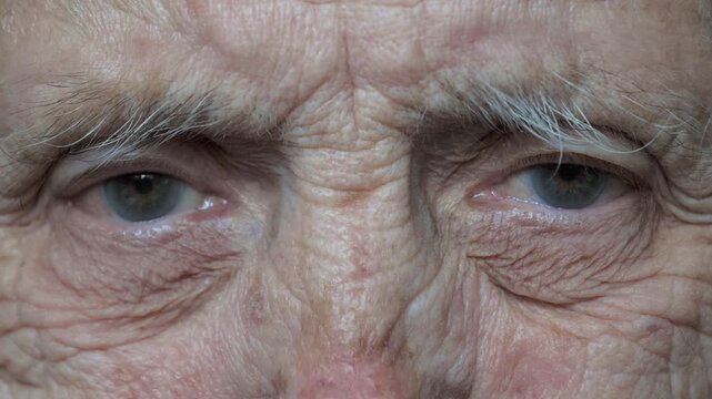 Extreme close up shot of 80 year old caucasian woman with deep wrinkles around blue eyes, blinking slowly and looking directly at camera, showing details of his aged and tired skin