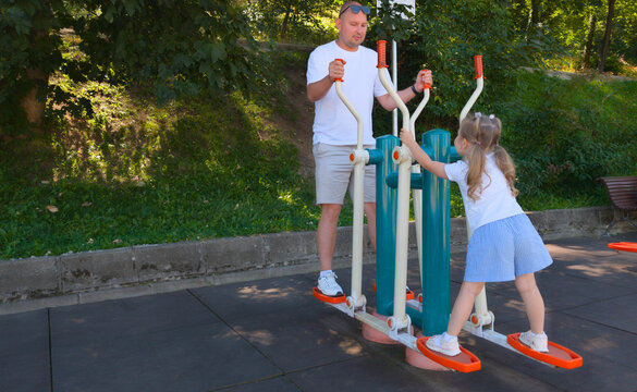 Happy Father And Little Daughter Exercising Together On Outdoor Elliptical Machine In Sunny Summer Park Ground